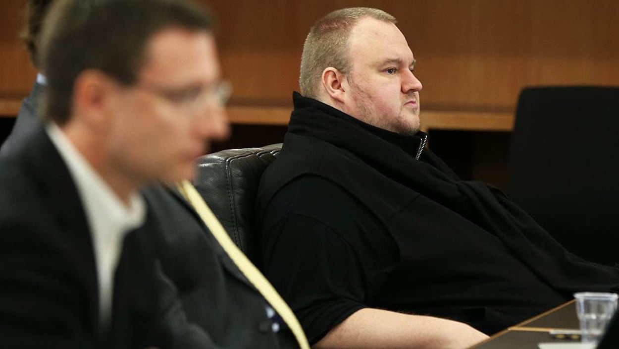 Kim Dotcom est assis dans une cour d'appel. L'air renfrogné, vêtu d'un gilet noir au col montant, il présente son profil droit et il a les cheveux courts.