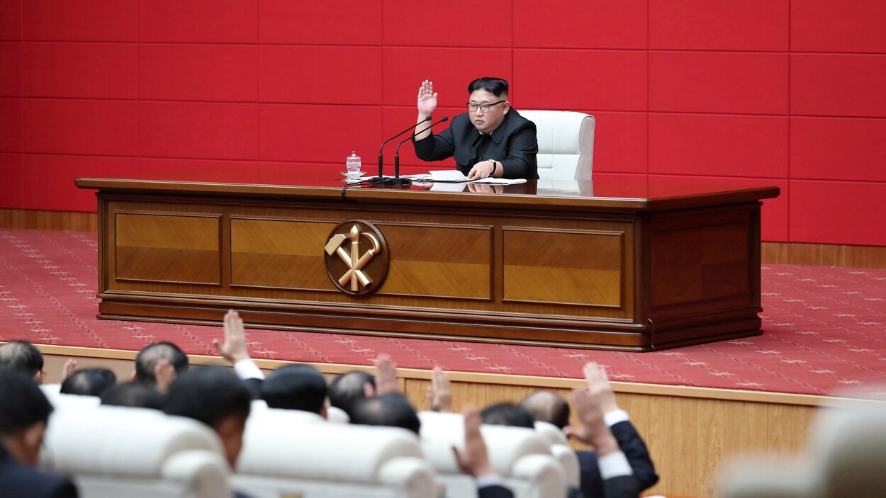 Kim Jong-un, assis seul derrière un massif bureau de bois, lève la main droite devant une assemblée d'hommes nord-coréens qui l'imitent.