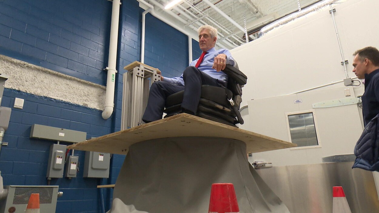 Un homme est assis sur une chaise perchée sur une plateforme de bois posée sur un robot à vibration caché par une bâche.