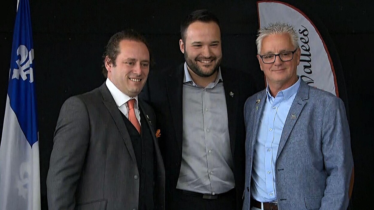 Les trois hommes, côte-à-côte, sourient sur l'estrade en regardant les photographes.