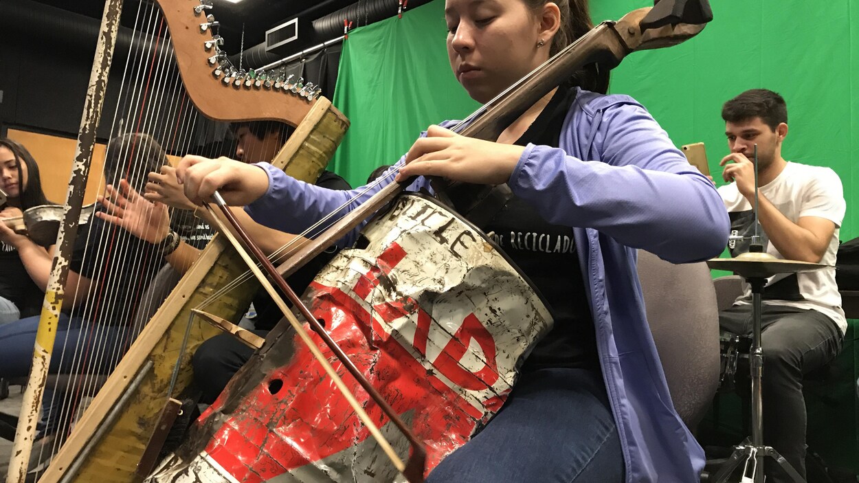 Une adolescente Paraguayen du Landfill Harmonic joue sur un violoncelle fabriqué à partir d'un bidon trouvé dans un dépotoir. Derrière elle un jeune homme joue de la harpe, elle aussi en matériau de récupération.