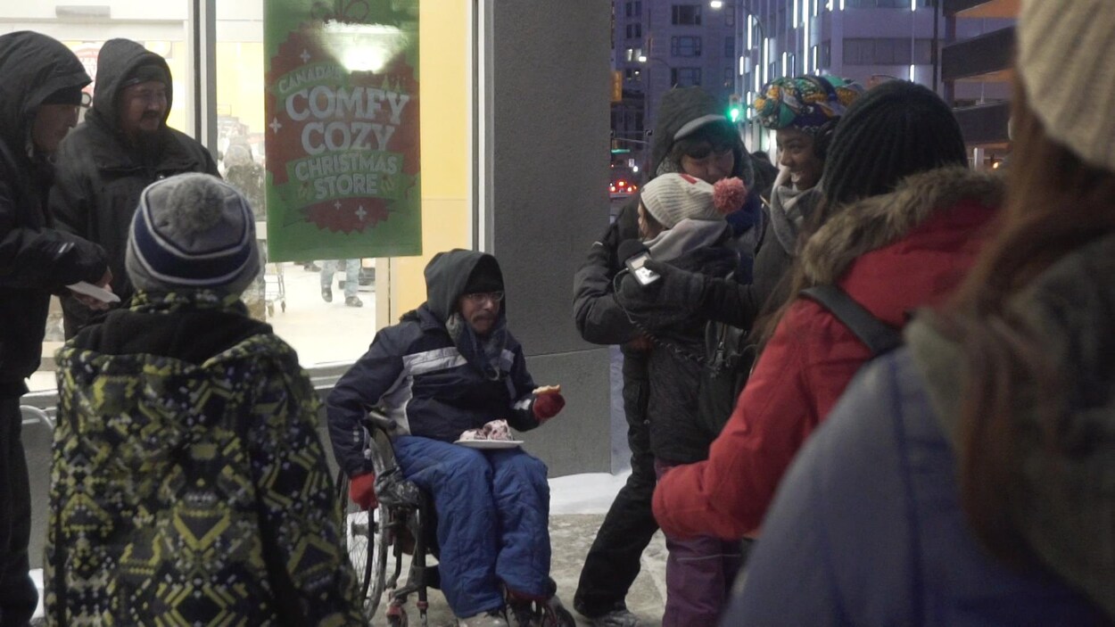 Un sans-abri en fauteuil roulant reçoit une pointe de pizza, entouré d'une demi-douzaine de personnes dans une rue enneigée de Winnipeg. Un sans-abri en fauteuil roulant reçoit une pointe de pizza, entouré d'une demi-douzaine de personnes dans une rue enneigée de Winnipeg.