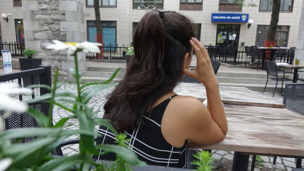 Une jeune femme aux cheveux foncés tourne le dos à la caméra en se cachant le visage, assise à une terrasse.