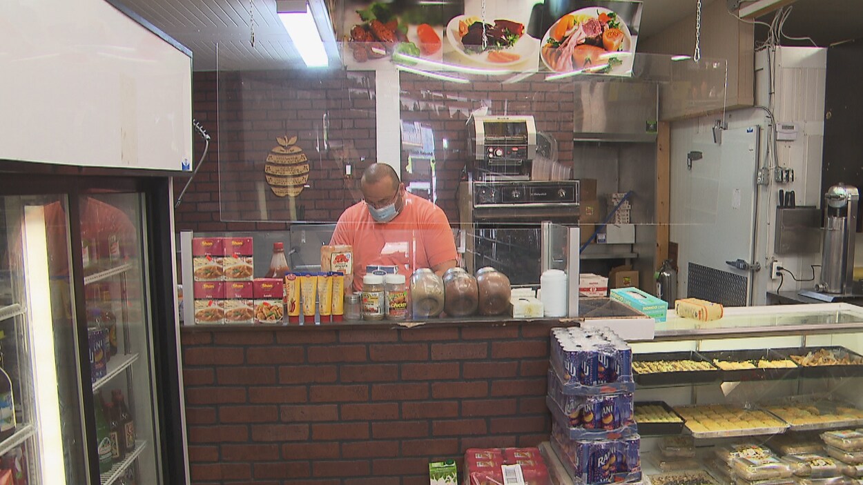 Un homme est derrière un comptoir dans un épicerie fine. Il porte un masque.