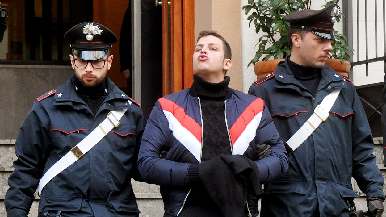 Un homme escorté par deux policiers envoie un baiser aux jounalistes.