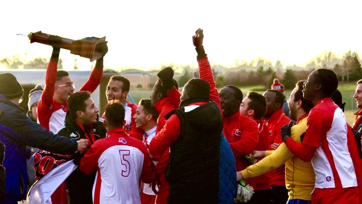 Des joueurs sont regroupés autour d'un des leurs qui portent bien haut le coupe du championnat qu'ls vienennt de remporter. On peut les voir rire et sourire et célébrer.