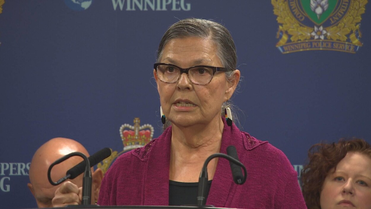 Une femme parle à deux microphones. Derrière elles sont assises deux autres personnes devant une bannière avec le logo de la police de Winnipeg. Une femme parle à deux microphones. Derrière elles sont assises deux autres personnes devant une bannière avec le logo de la police de Winnipeg.