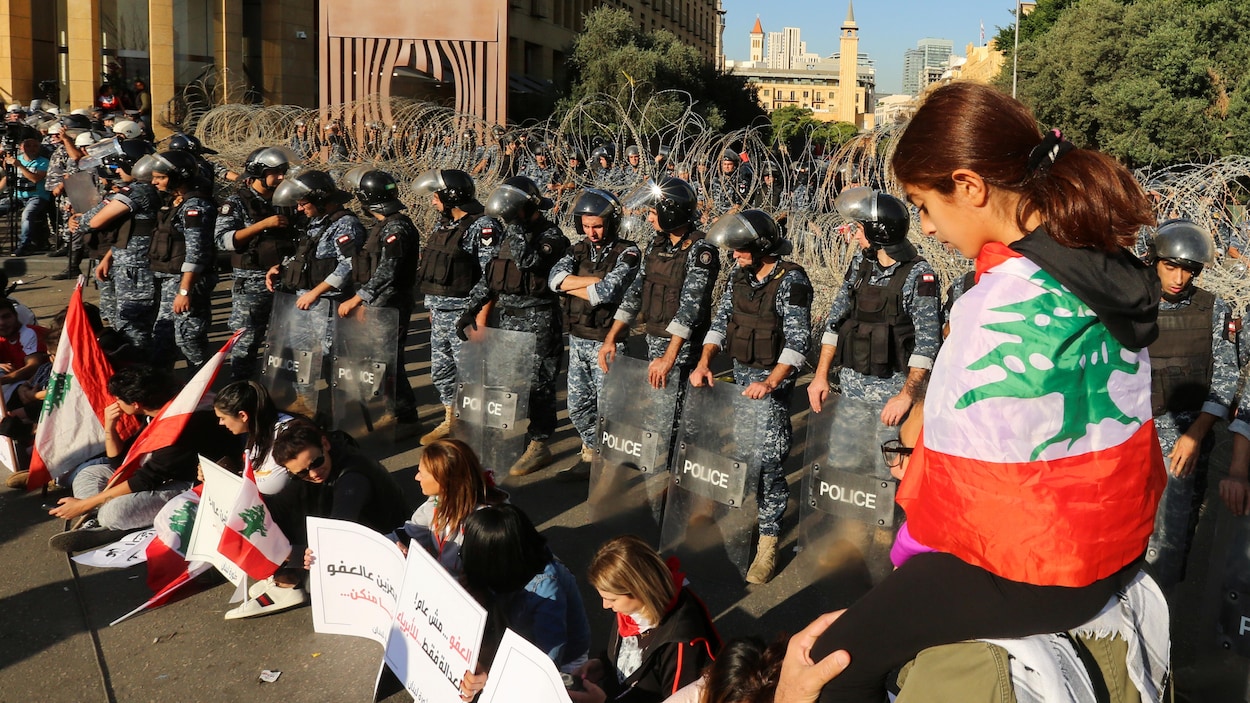 Une fillette qu'un homme porte sur ses épaules est drapée du drapeau libanais. Devant elle, des femmes tenant des pancartes sont assises devant des policiers antiémeute.
