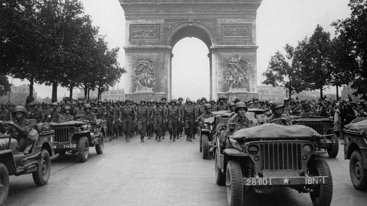 Photographie en noir et blanc d'une parade militaire sous l'Arc de Triomphe avec des soldats et des voitures.