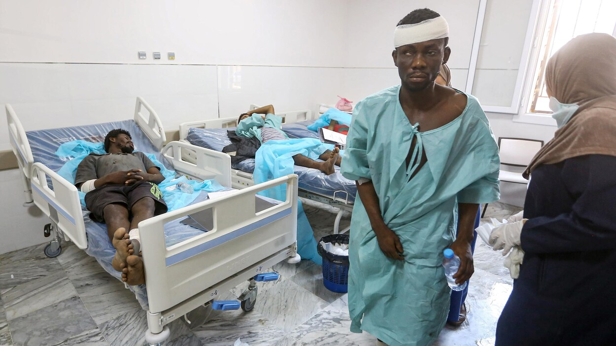 Un homme marche en jaquette d'hôpital portant des bandages à la tête et à une cheville. Deux autres personnes sont couchées dans des lits.