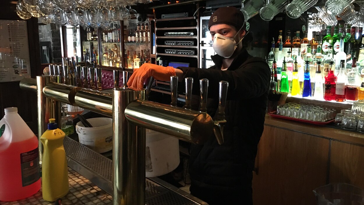 Un homme avec un masque et des gants active un fût de bière dans un bar.