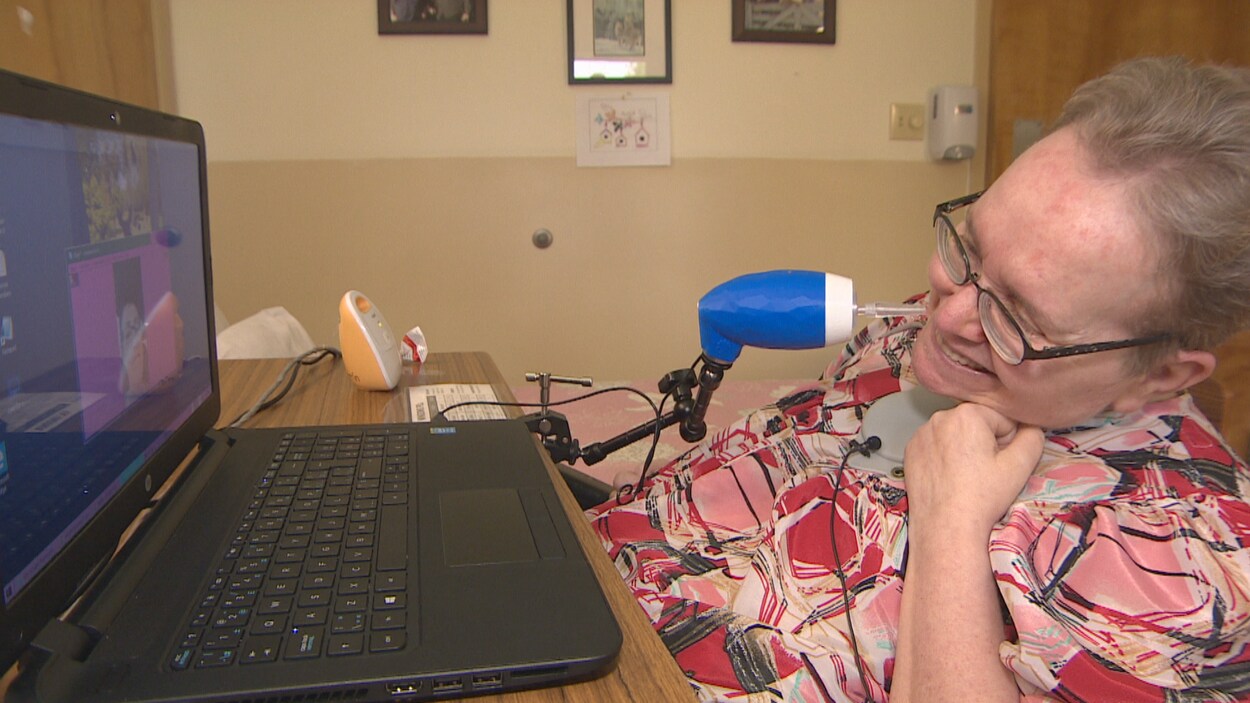 En femme quadriplégique regarde son ordinateur en souriant, un appareil ovale, monté sur un pied articulé et avec un embout se trouve proche de sa bouche.