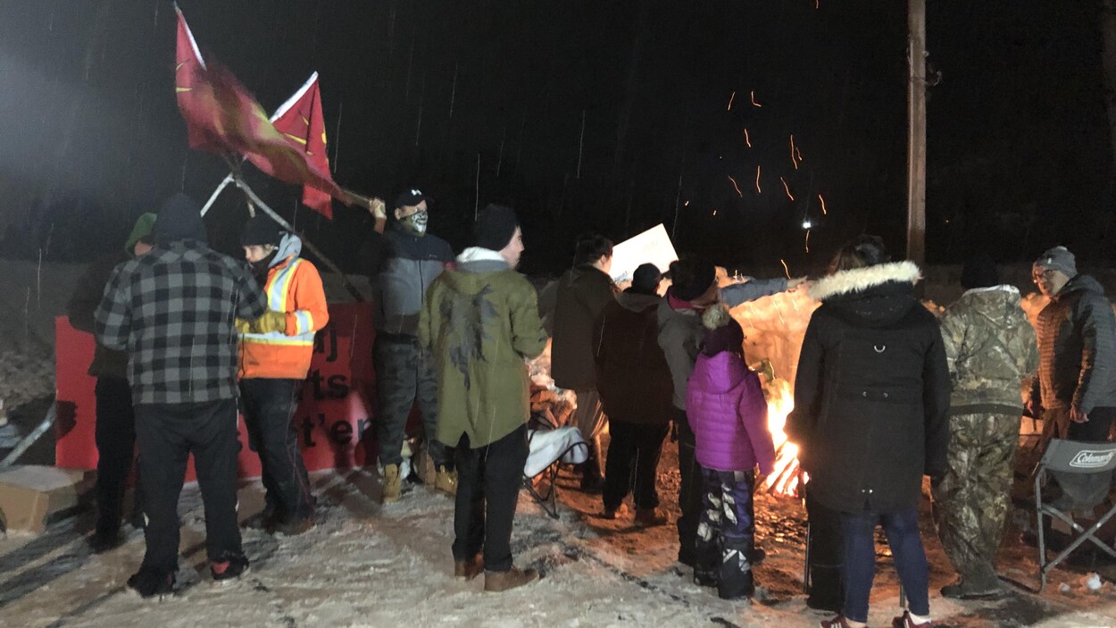 Une douzaine de personnes sont debout sur les rails enneigés et se réchauffent près d'un feu.