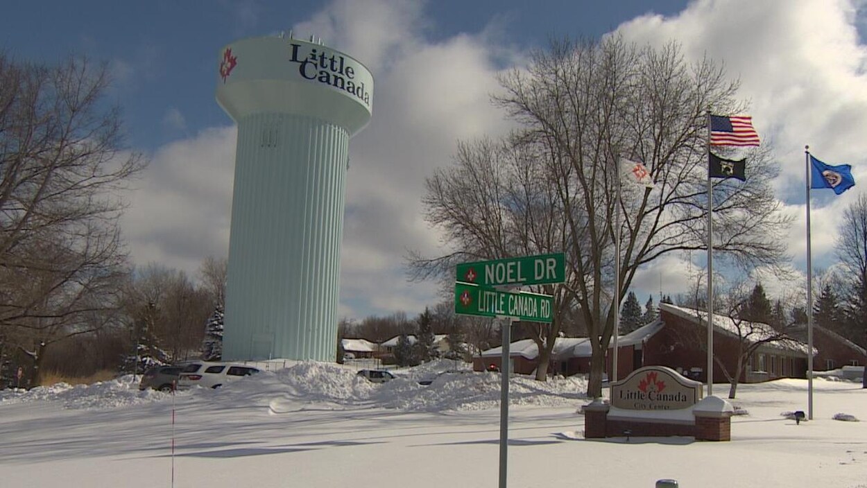 Une tour d'eau et un panneau près d'un drapeau américain aux abords d'une route enneigée portent la mention Little Canada et le logo d'une feuille d'érable. Une tour d'eau et un panneau près d'un drapeau américain aux abords d'une route enneigée portent la mention Little Canada et le logo d'une feuille d'érable.