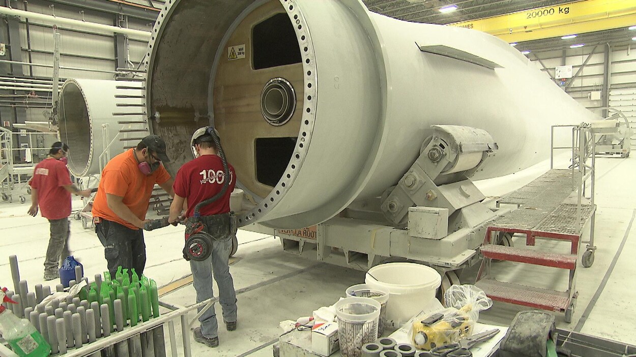 Trois hommes s'affairent dans une usine, près d'une éolienne en construction.