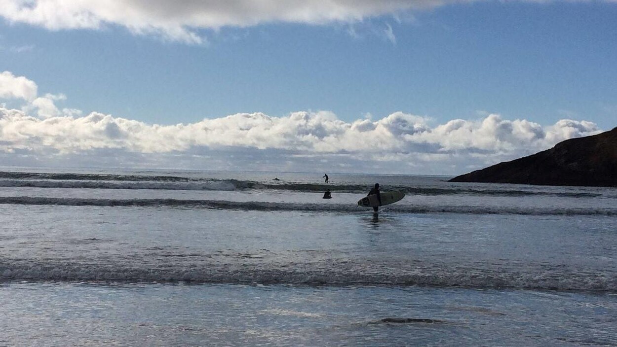 Un homme tient sa planche de surf debout dans l'eau alors que des vagues déferlent sur le rivage. Deux autres surfeurs au loin, un debout, un assis sur sa planche.