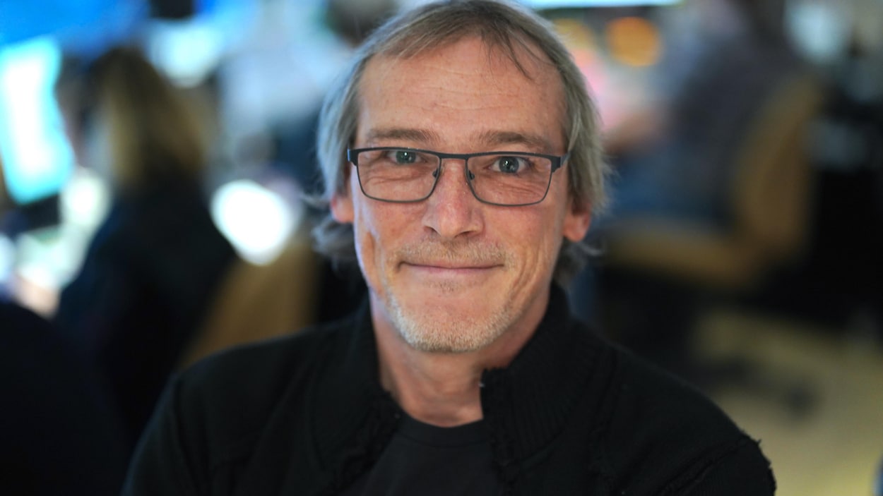 Un homme avec des lunettes regarde vers la lentille de l'appareil photo et sourit.