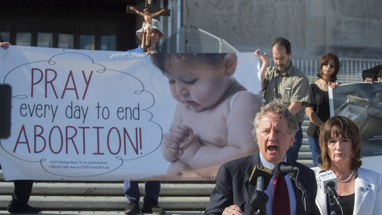John Milkovich et Valarie Hodges s'adressent aux médias devant une affiche montrant un bébé à côté des mots « Priez chaque jour pour mettre fin à l'avortement », au-dessus de laquelle se trouve un crucifix.