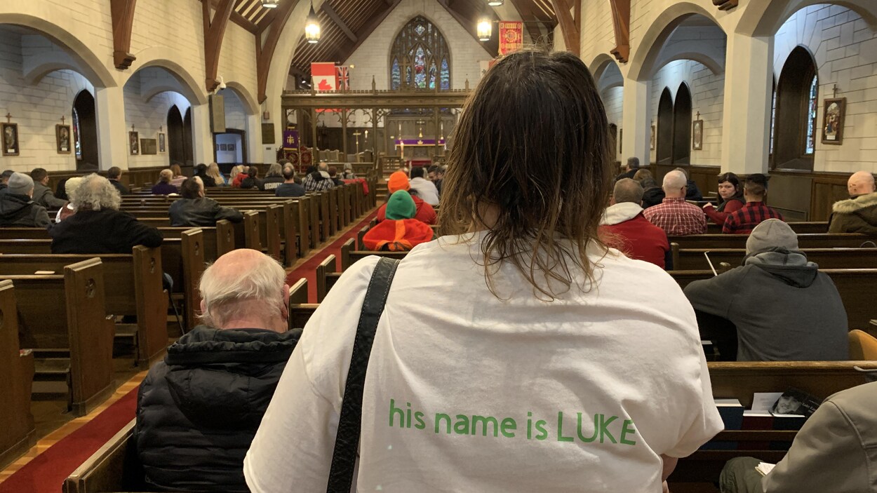 Une femme de dos, debout dans une église. Les mots « Son nom est Luke » sont imprimés en vert dans le dos de son chandail blanc. D'autres personnes sont assises sur les bancs de l'église.