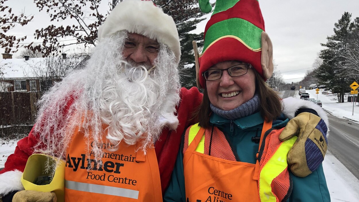 Deux bénévoles, l'un habillé en père Noël et l'autre en lutin, se tiennent côte à côté à l'extérieur en souriant.