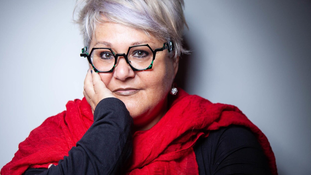 Portrait en couleur de l'autrice Lynda Dion, une main sur la joue droite, foulard rouge autour du cou, lunettes vertes hexagonales, cheveux courts.