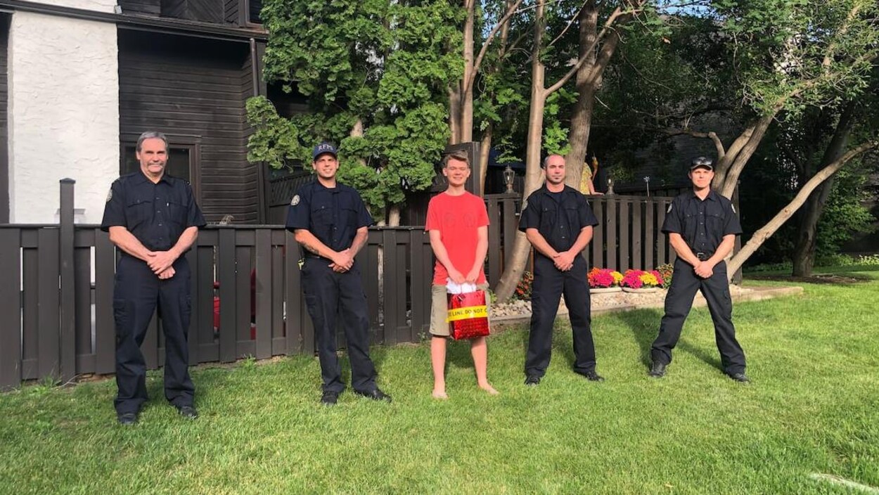 Mac Findlay est debout à l'extérieur. De chaque côté de lui se trouvent des pompiers en uniforme.
