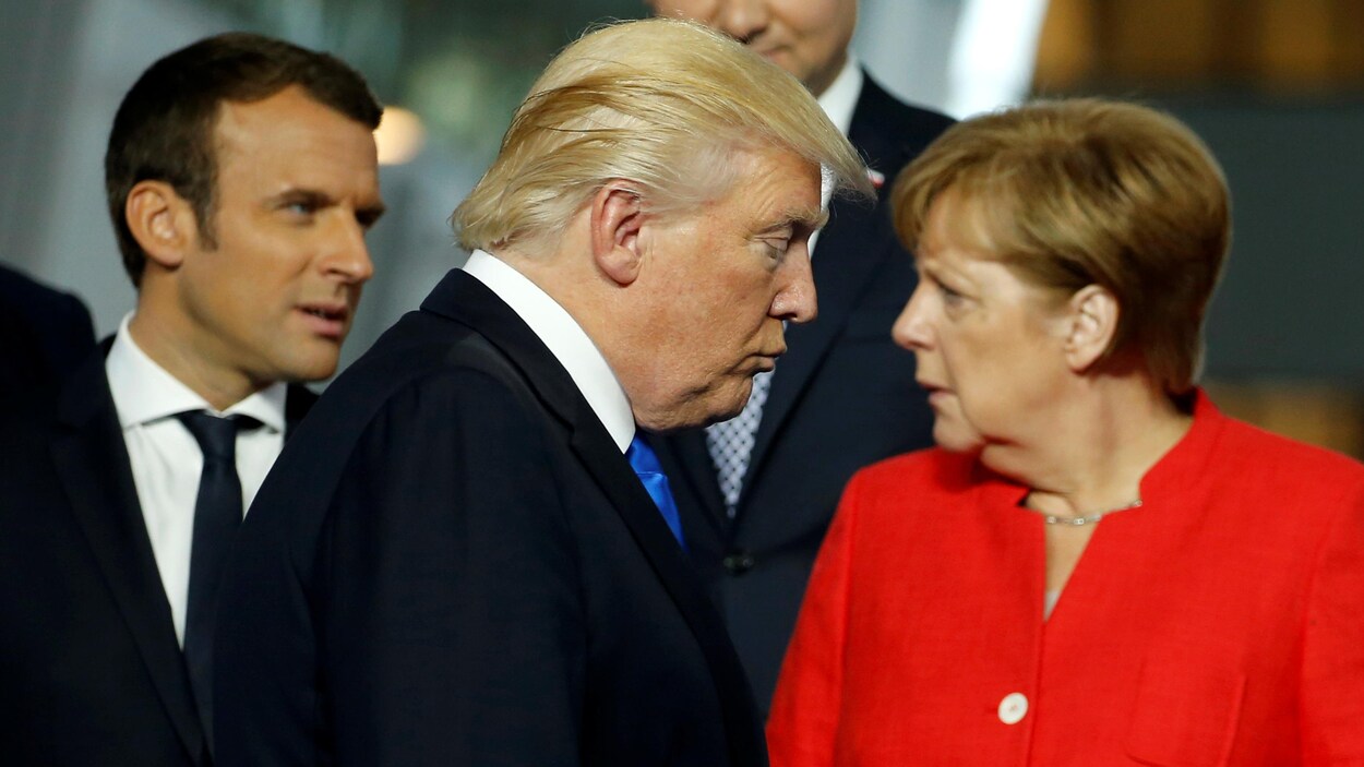 Donald Trump regarde vers le sol tandis qu'Emmanuel Macron et Angela Merkel discutent derrière lui.
