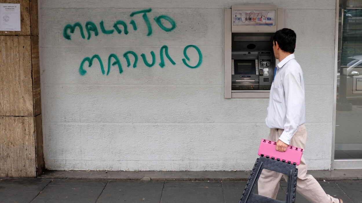 Un homme passe devant un guichet automatique près duquel un graffiti dit : « Allez au diable Maduro! ».