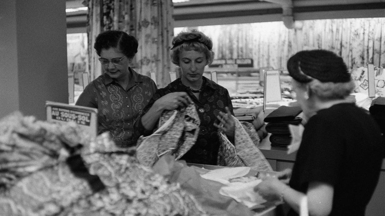 En noir et blanc, trois femmes manipulant des vêtements dans un rayon du magasin Dupuis Frères En noir et blanc, trois femmes manipulant des vêtements dans un rayon du magasin Dupuis Frères