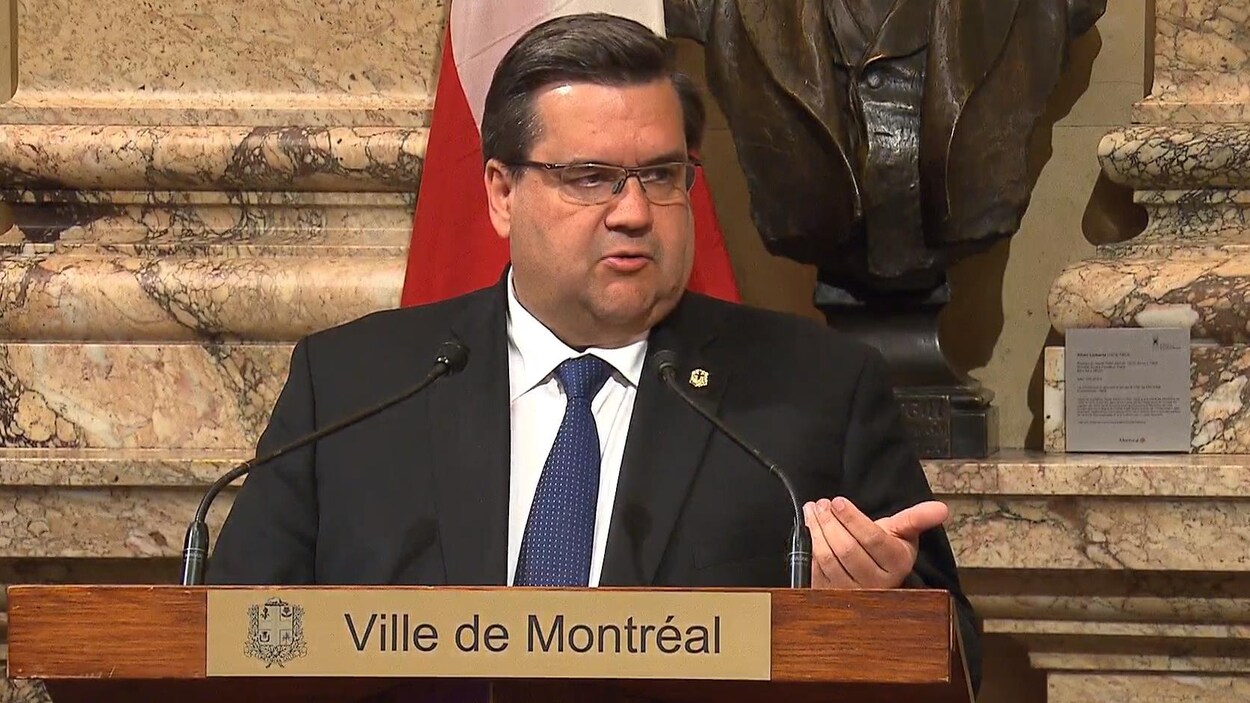 Le maire Denis Coderre en conférence de presse à l'hôtel de ville de Montréal, au podium, avec une statue à l'arrière et un drapeau.