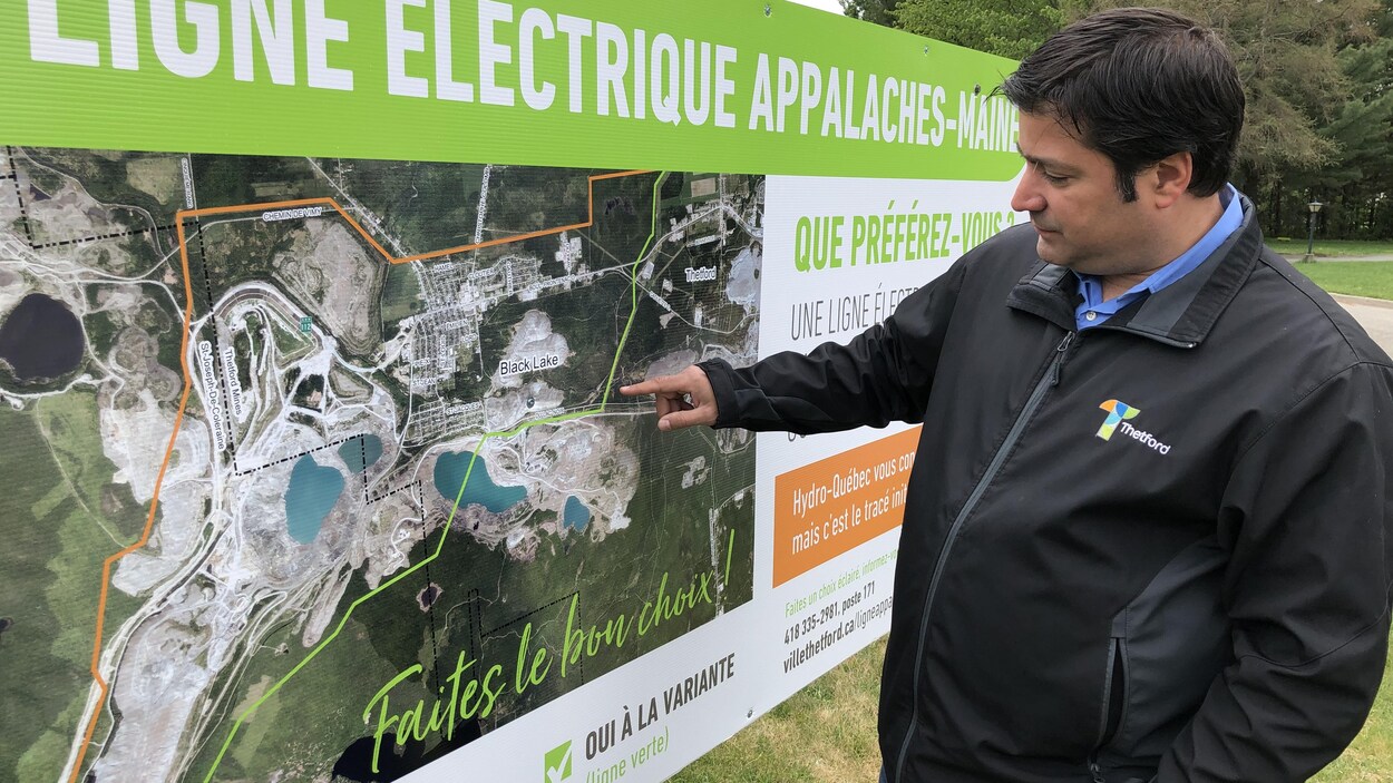 Le maire debout devant une affiche présentant les tracés proposés pour la ligne électrique Appalaches-Maine d'Hydro-Québec.