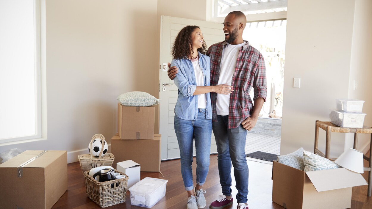 Un jeune couple est debout à l'intérieur d'une maison avec des boîtes autour d'eux.