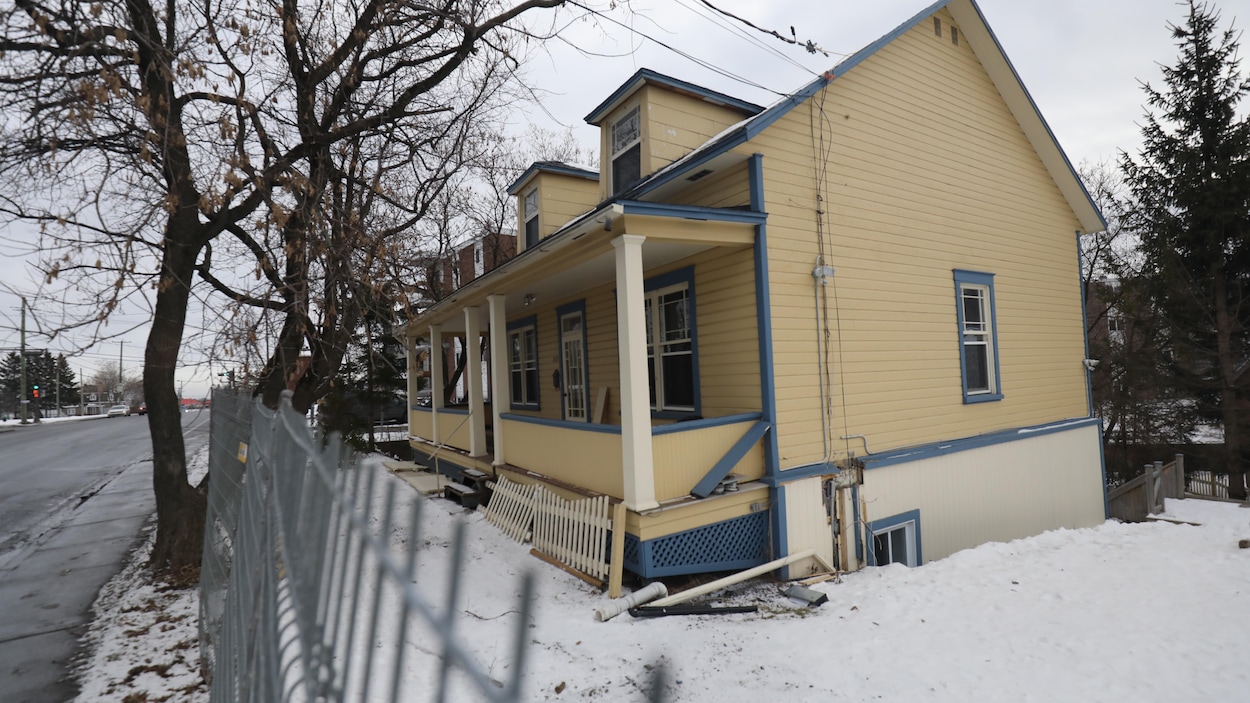 Une maison jaune et bleue, avec une petite galerie en avant, et des clôtures de construction, sur un terrain enneigé.