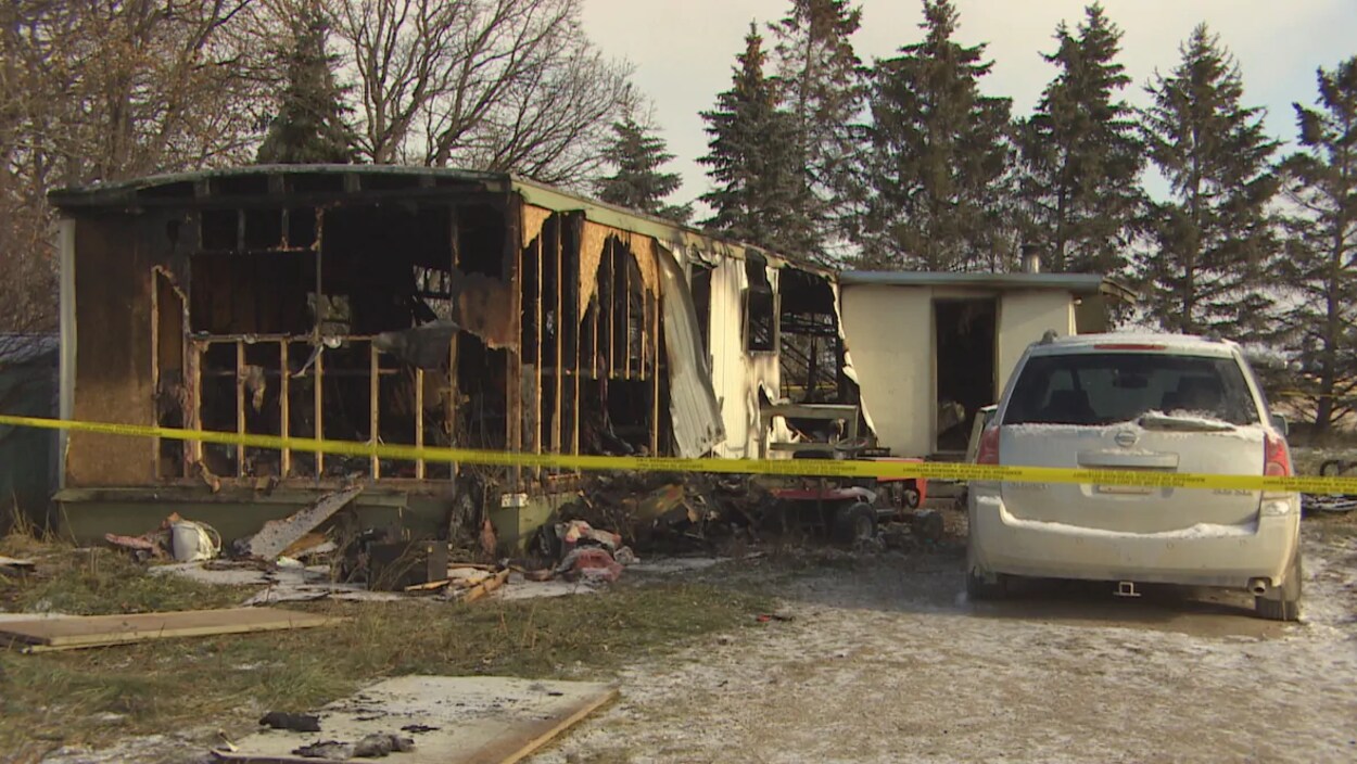 Une voiture est garée à côté de la maison modulaire détruite par le feu et qui est entourée par un ruban jaune de la police.