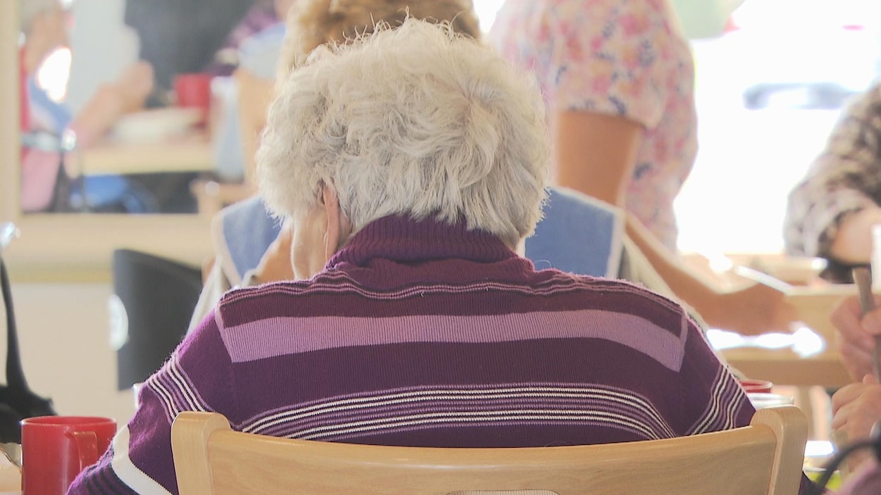 Personnes âgées assises à des tables et une préposée aux bénéficiaires qui sert de la nourriture. Personnes âgées assises à des tables et une préposée aux bénéficiaires qui sert de la nourriture.