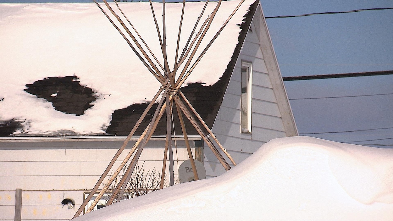 Derrière un gros banc de neige, de longues branches forment un tipi, tout près d'une maison.