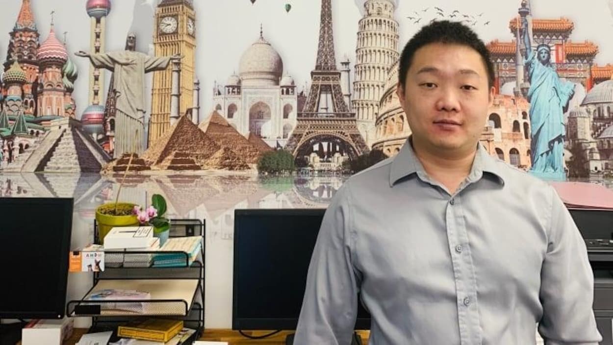 Un homme debout dans une salle. Derrière lui, une affiche qui montre les monuments de plusieurs capitales dans le monde.