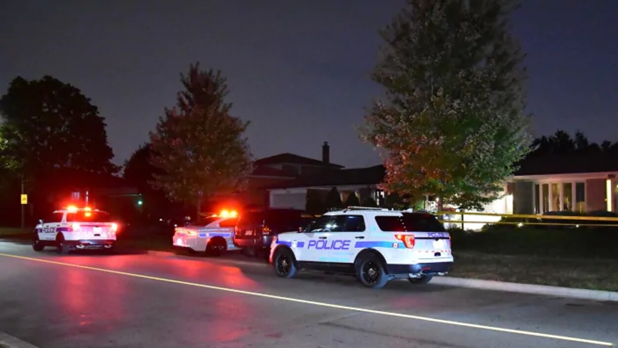 Photo de trois véhicules de police le soir garés devant une résidence dans une rue; leurs gyrophares sont allumés.