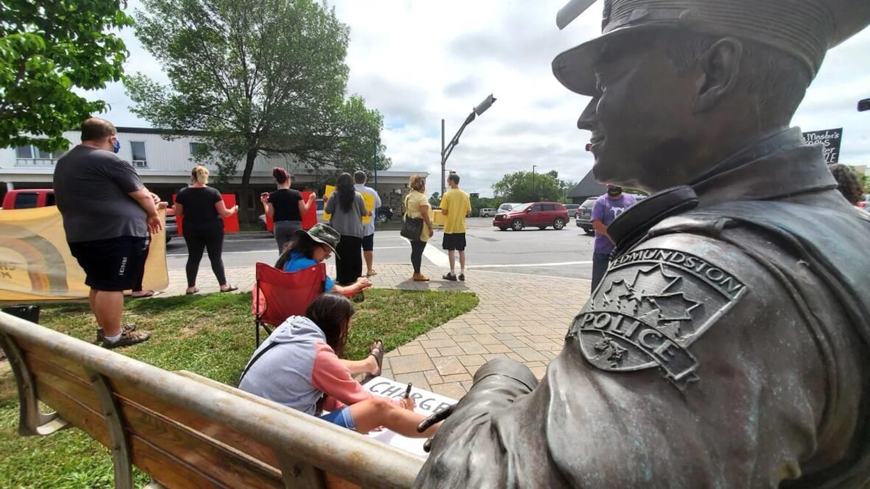 Une statue représentant un policier assis sur un banc. Des manifestants sont debout près de cet endroit. Une femme assise par terre dessine une affiche pour la manifestation.