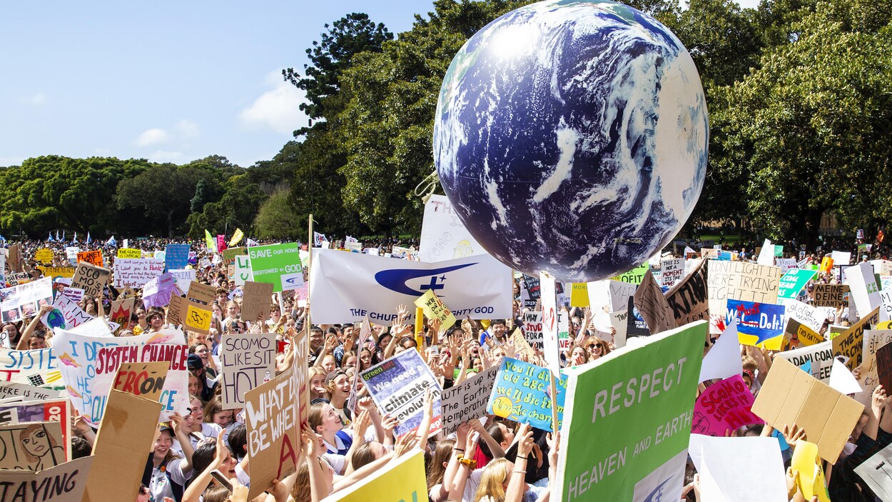 Des centaines de manifestants, en Australie, tiennent toutes sortes d'affiches et un grand ballon représentant la Terre.