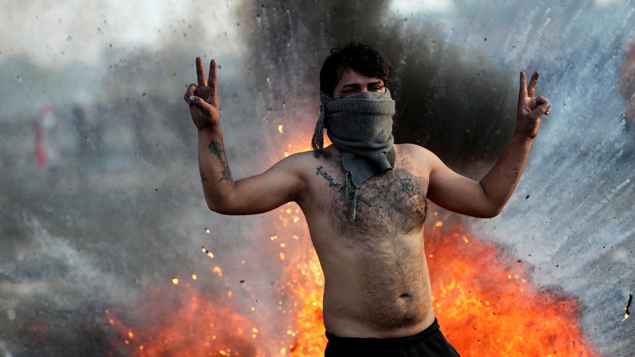 Un homme torse nu faisant le signe de la victoire avec ses deux mains avec en arrière-plan un feu.