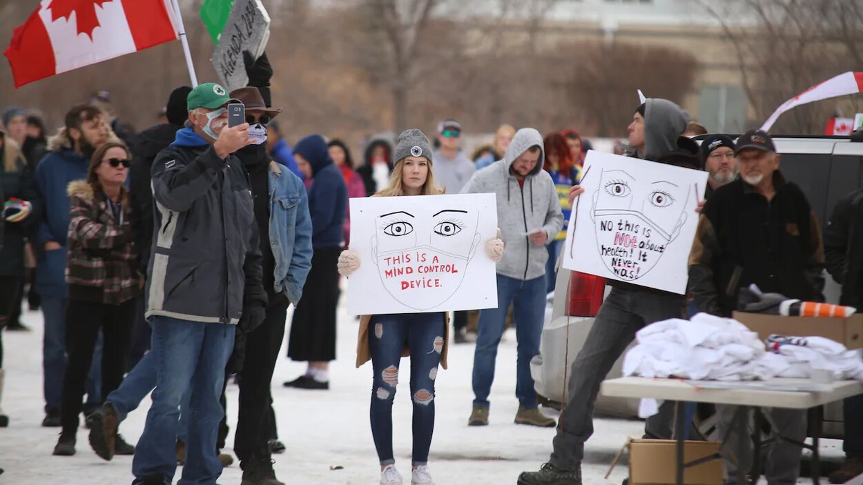 Des manifestants sont regroupés, deux d'entre eux tiennent des pancartes disant que le port du masque représente un contrôle.