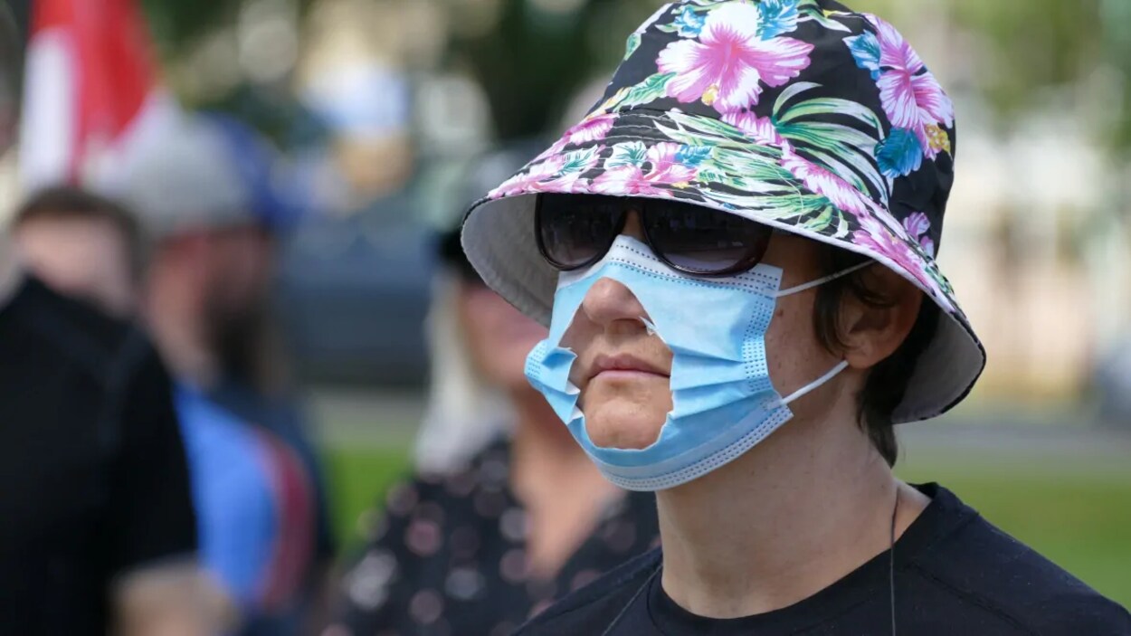Une femme portant un chapeau, des lunettes de soleil et un masque avec un trou découpé au niveau de la bouche.