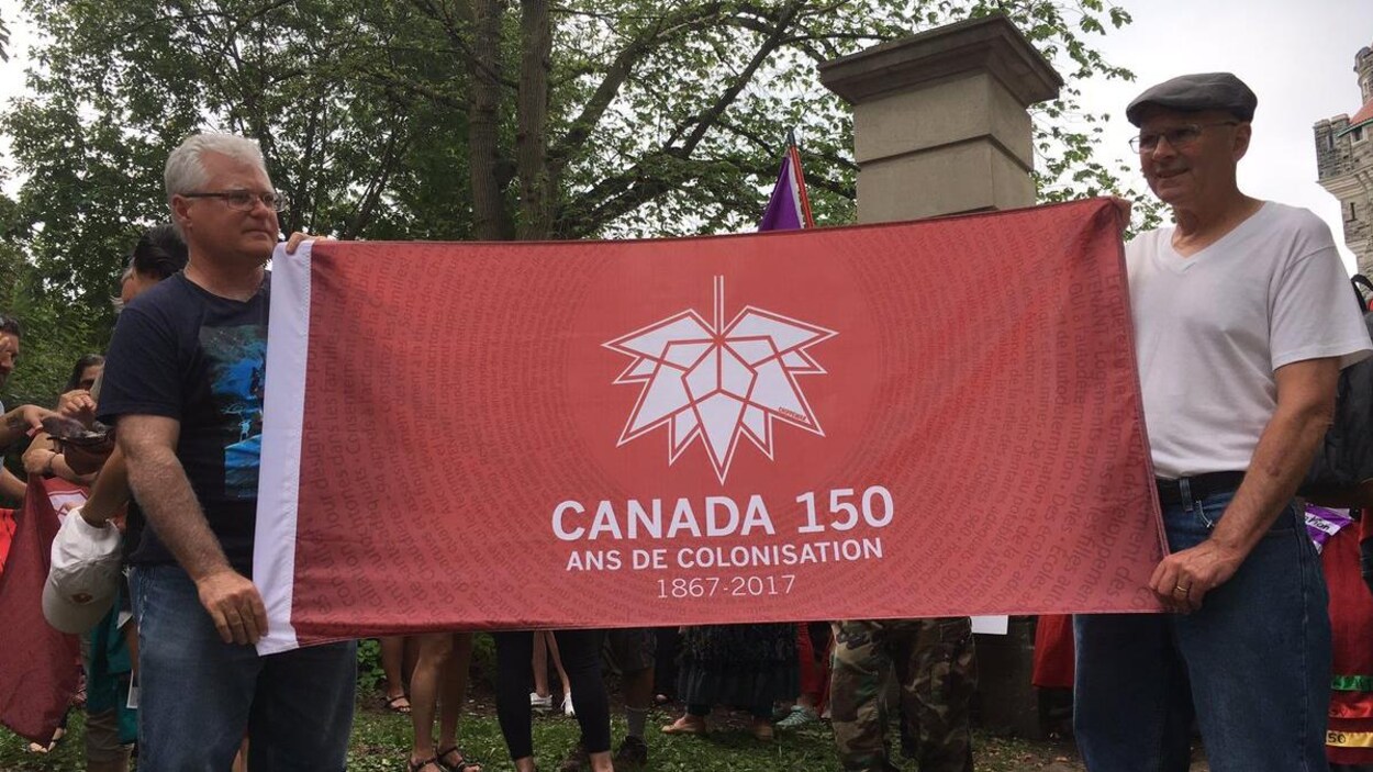 Deux hommes tiennent un drapeau rouge où il est écrit «Canada 150 ans de colonisation » Deux hommes tiennent un drapeau rouge où il est écrit «Canada 150 ans de colonisation »