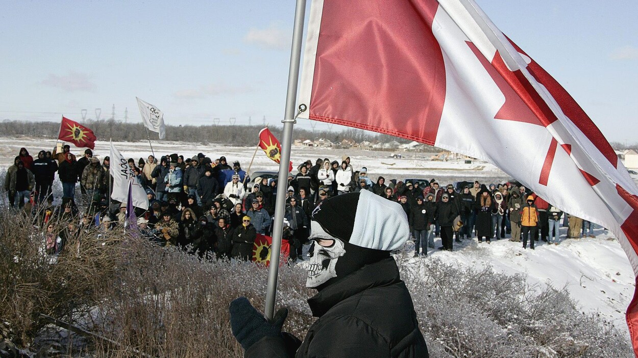 Une personne masquée porte un drapeau du Canada devant un groupe d'environ 150 Autochtones.