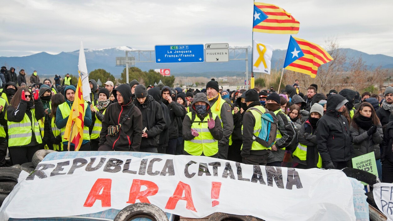 Plusieurs centaines de personnes bloquent une autoroute. Certains sont vêtus de noir ou de dossards jaunes. On voit le drapeau catalan qui flotte dans la foule.