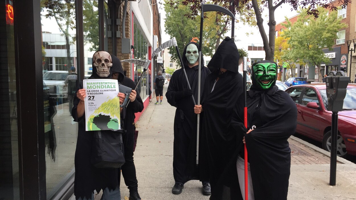 Cinq personnes tiennent une faux, sont habillées en noir et portent un masque.