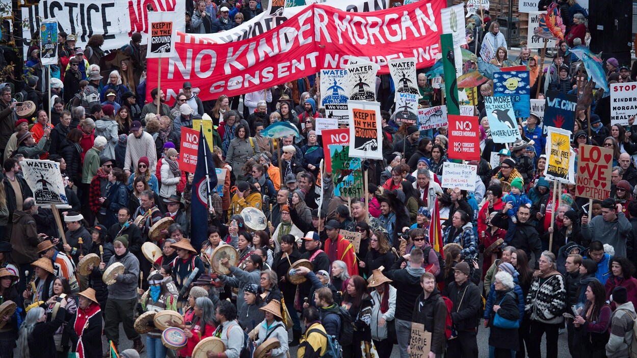 Des milliers de manifestants, certains en habits traditionnels autochtones, et plusieurs qui tiennent des pancartes sur lesquelles on peut lire an anglais Non Kinder Morgan, sont rassemblés.