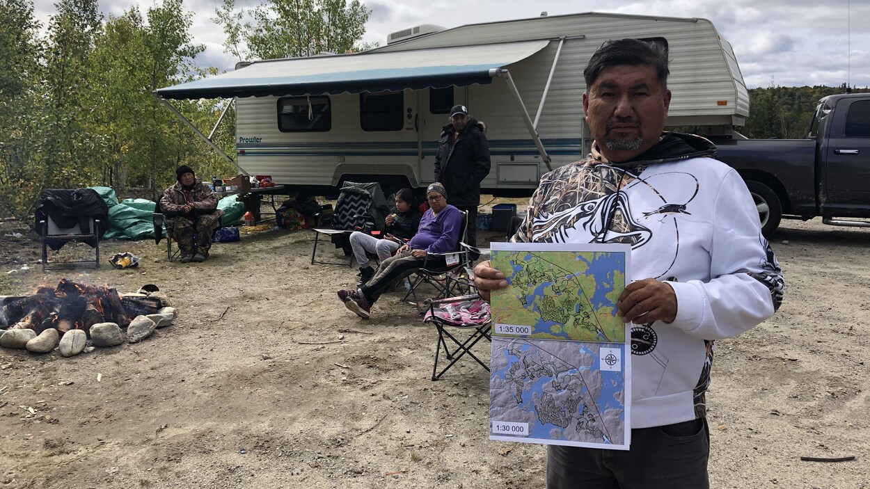 Edmond Brazeau tient une carte dans ses mains. D'autres personnes derrière lui sont assises dans des chaises de camping à côté d'une roulotte.