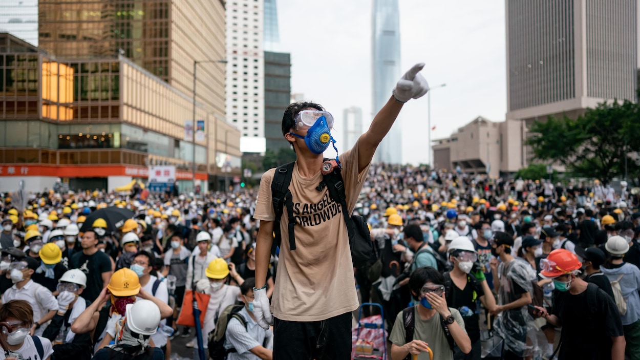 Un jeune portant un masque se tient debout, un bras levé, devant une foule de manifestants.
